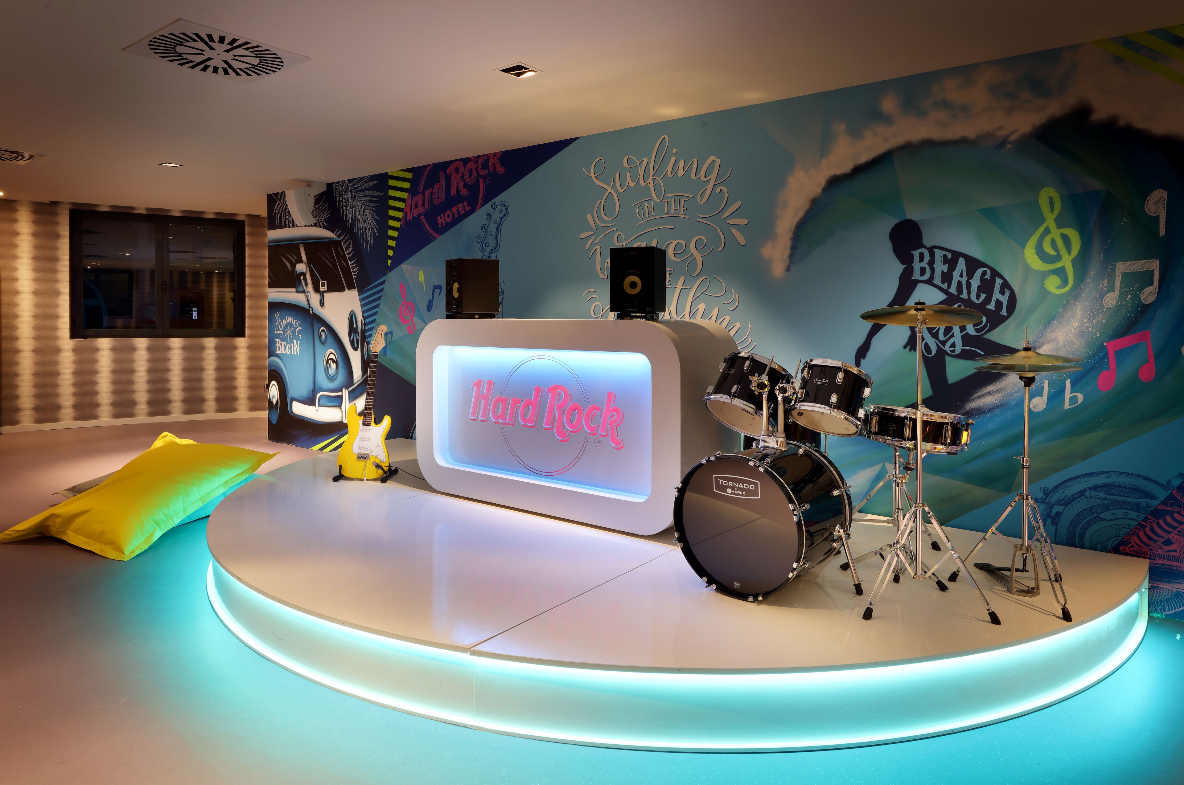 A stage with a drum set and a neon sign reading "Hard Rock" in a room with a mural of a car and musical notes.