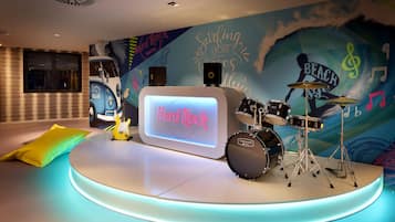 A stage with a drum set and a neon sign reading "Hard Rock" in a room with a mural of a car and musical notes.