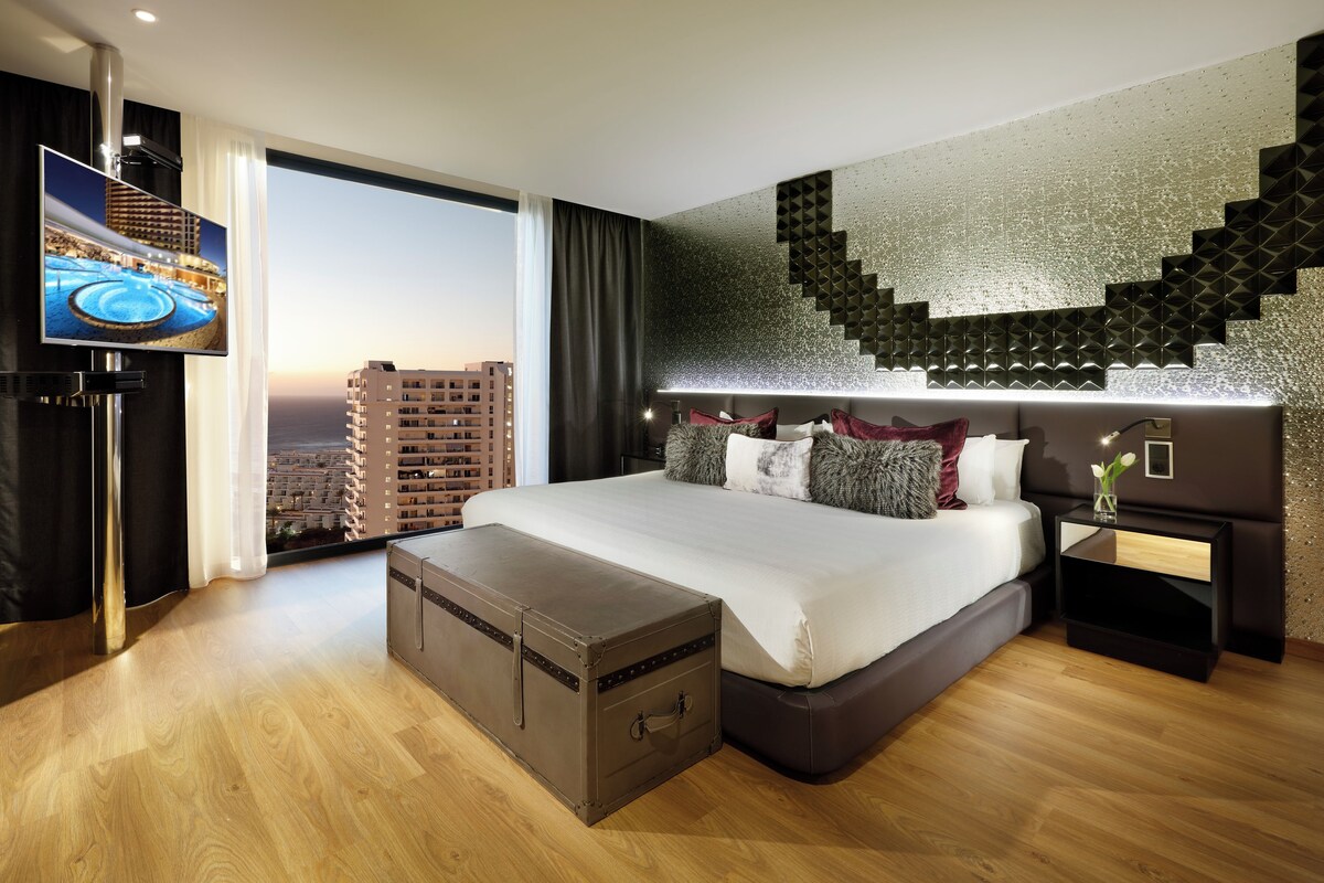 Una habitación de hotel moderna con una cama grande, un televisor, una maleta y vistas a la ciudad.