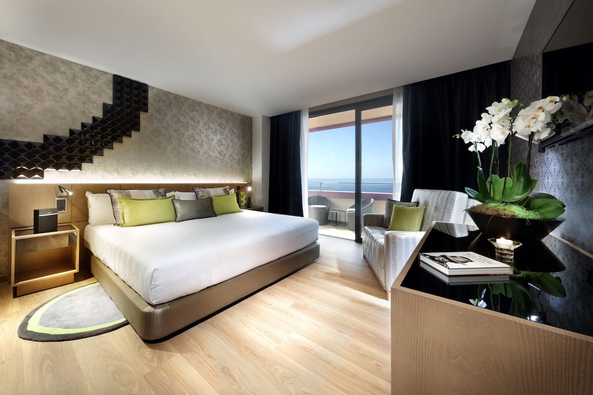 Moderni hotellihuone, jossa on suuri sänky, portaikko, parvekkeelta avautuva näköala ja pieni pöytä, jonka päällä on kasvi.
