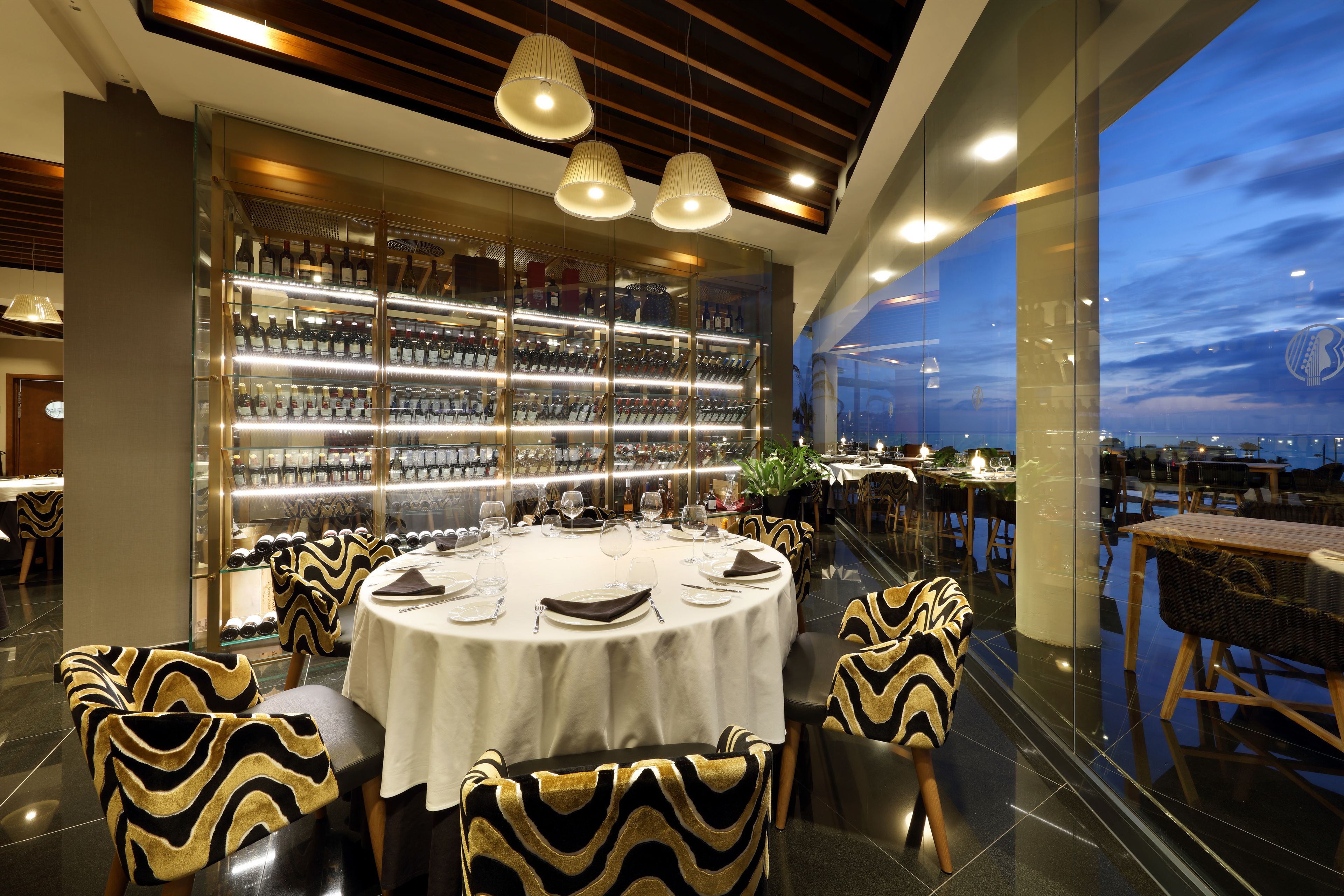 A modern restaurant with a glass wall offering a city view, a well-lit bar area, and elegantly designed seating.