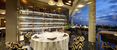 A modern restaurant with a glass wall offering a city view, a well-lit bar area, and elegantly designed seating.