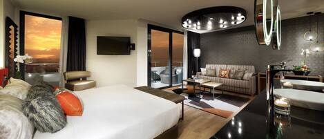 A modern hotel room with a large bed, a sitting area with a sofa and coffee table, a bathroom with a vanity and mirror, and a city view through large windows.