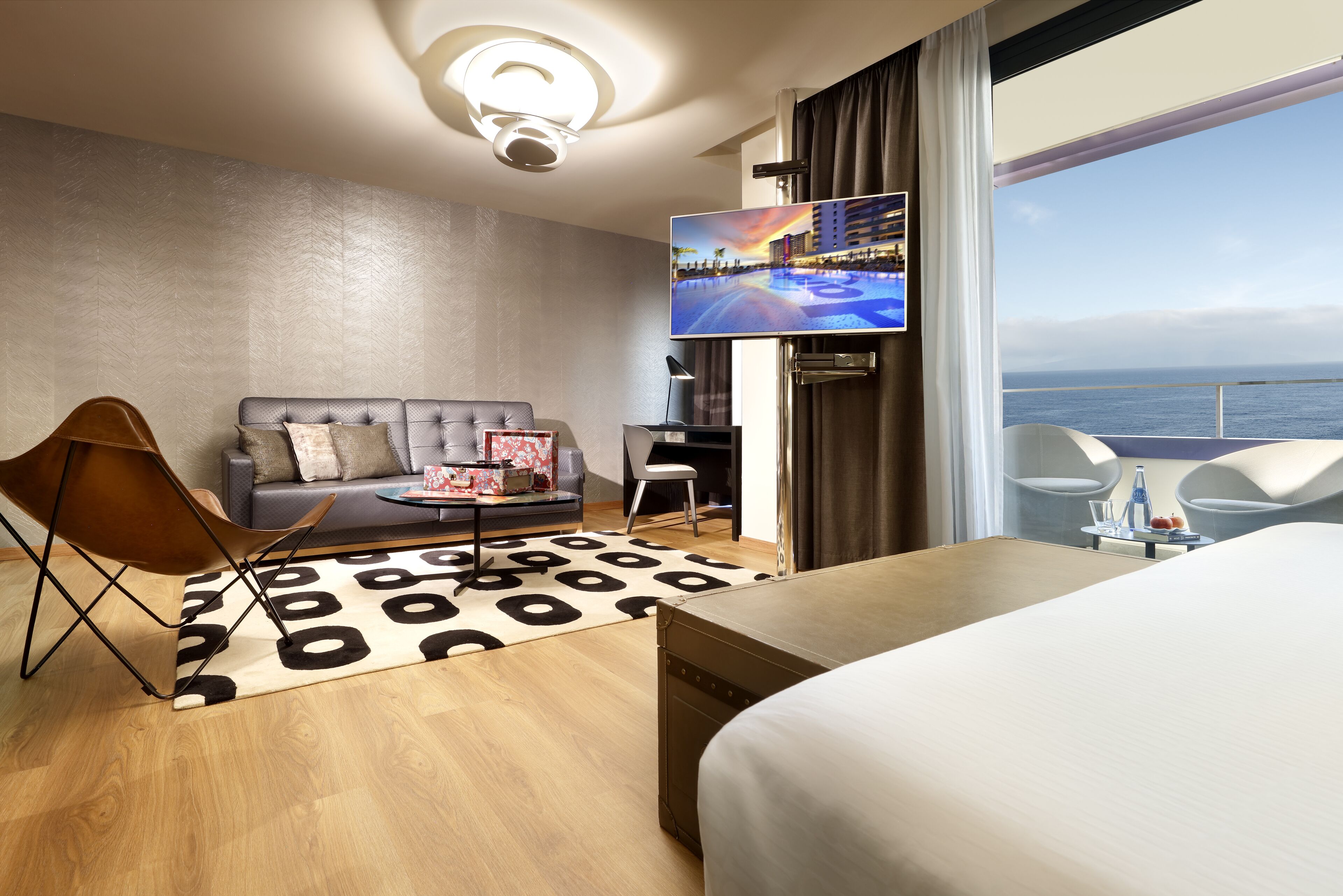 A modern hotel room with a large bed, a sofa, a TV, and a view of the sea.