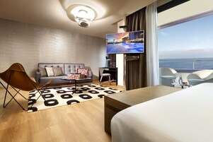 A modern hotel room with a large bed, a sofa, a TV, and a view of the sea.
