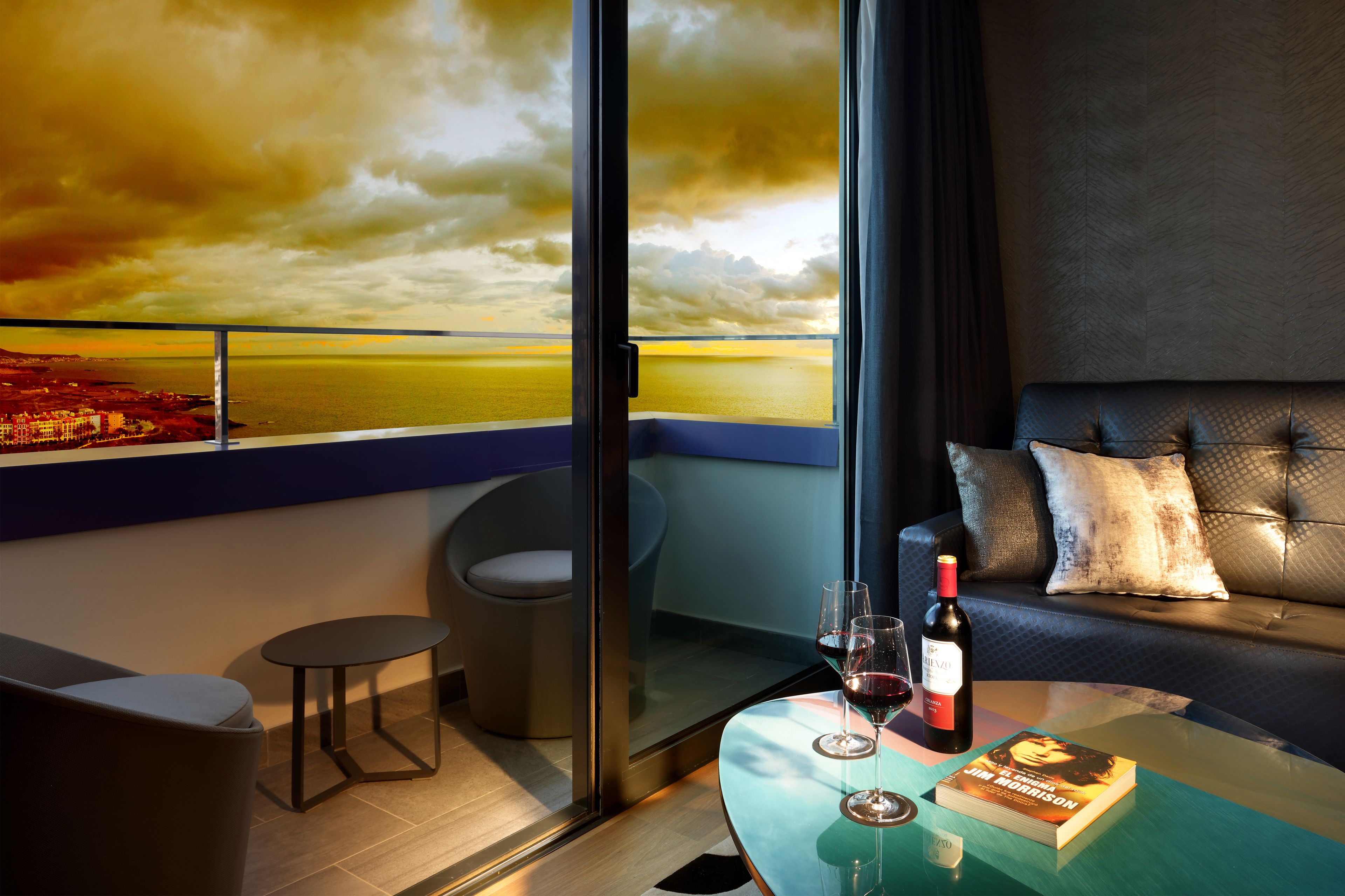 A modern room with a glass table, a sofa, and a view of a sunset over the sea.