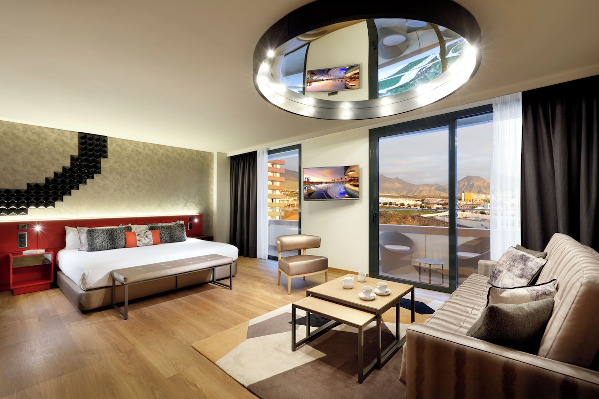Una moderna camera d'albergo con un letto, un divano, un tavolino da caffè e un'ampia finestra che si affaccia sulla città.