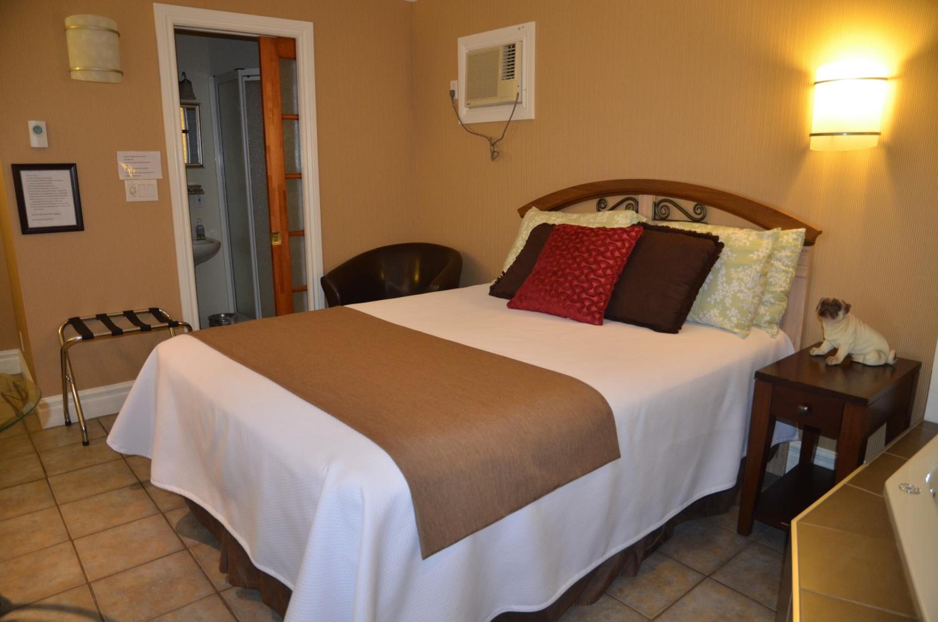 A hotel room with a large bed, a bedside table, a chair, a small table, a lamp, and a wall-mounted air conditioner.