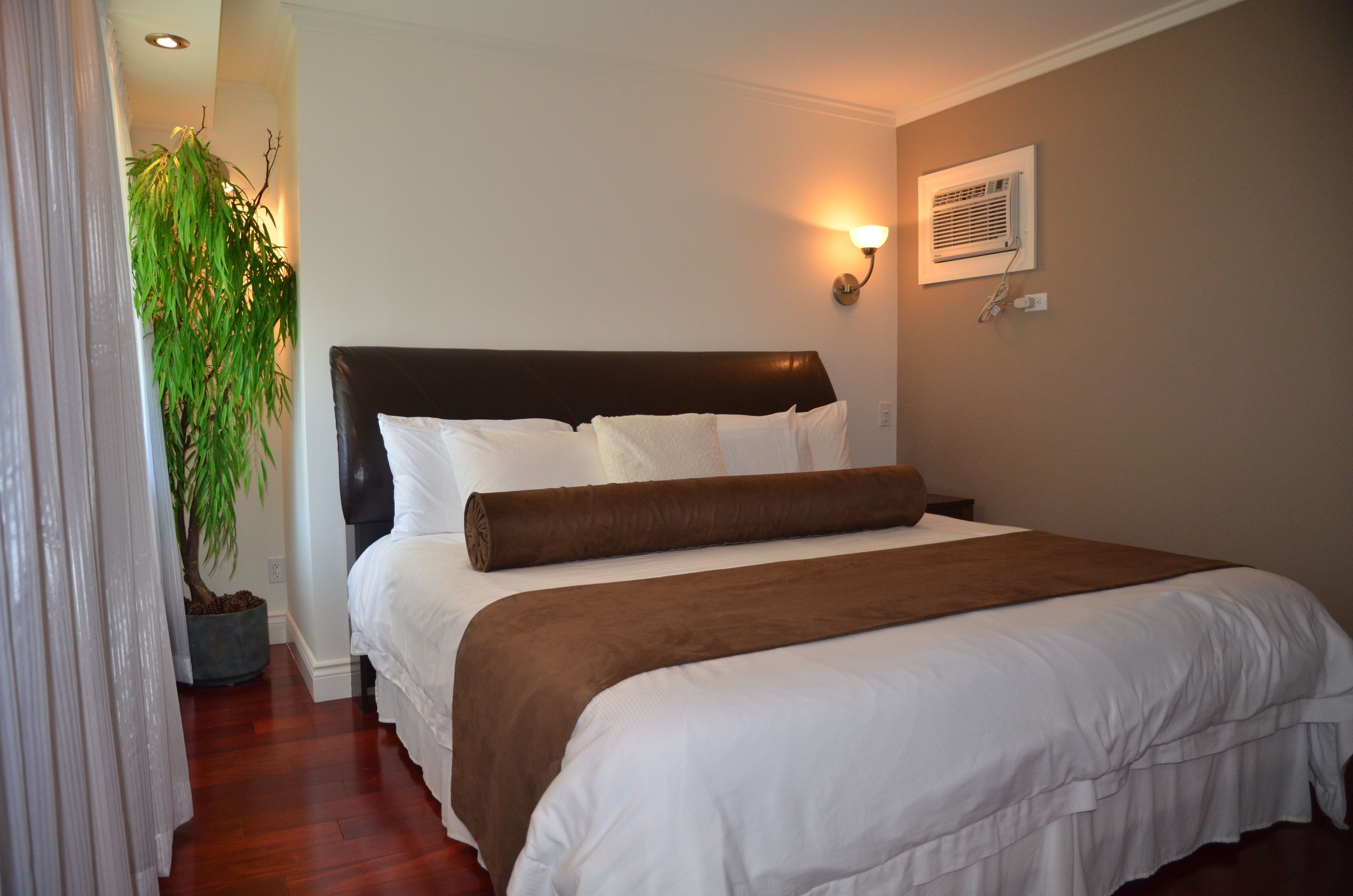 Una cama bien hecha con ropa de cama blanca y marrón, una planta en maceta y un aire acondicionado de pared.