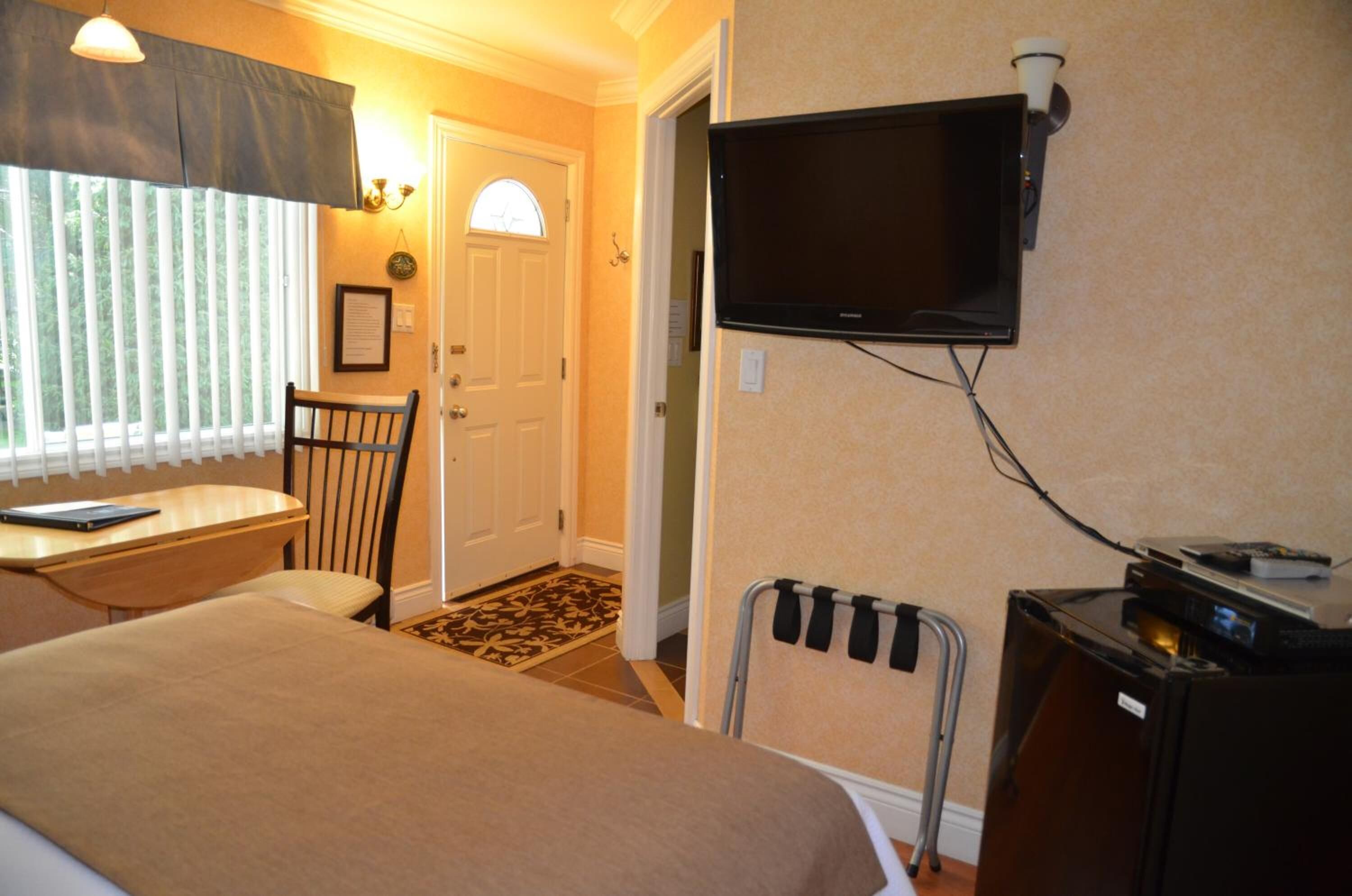 A hotel room with a bed, a desk with a chair, a television mounted on the wall, and a small refrigerator.