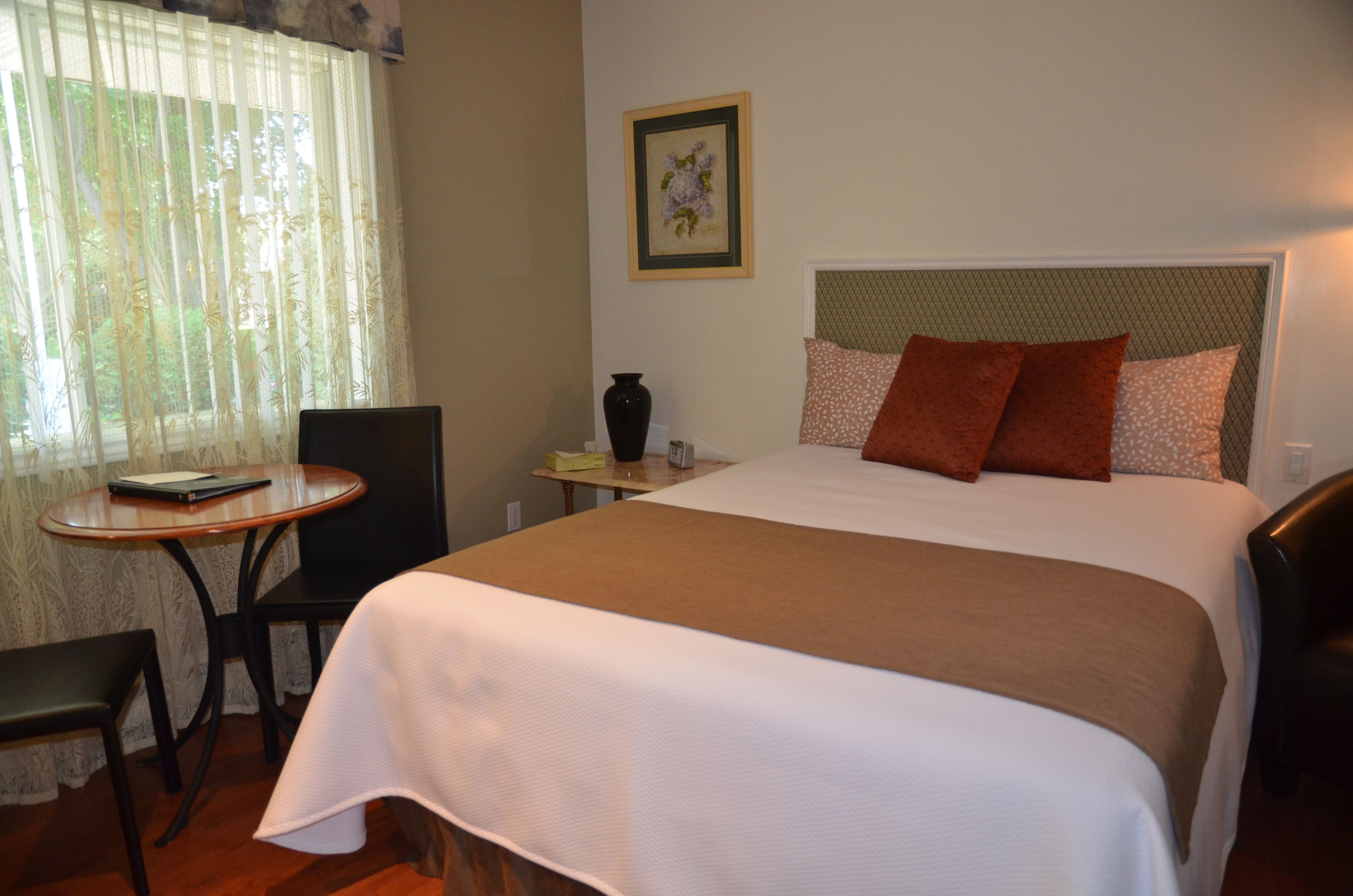 A hotel room with a bed, a desk, a chair, a vase, and a framed picture on the wall.