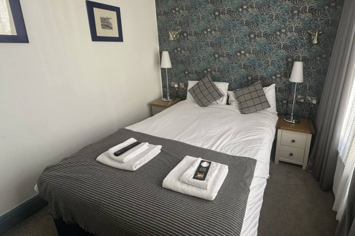 A hotel room with a bed, two bedside tables, a nightstand, a window with curtains, and a patterned wallpaper.