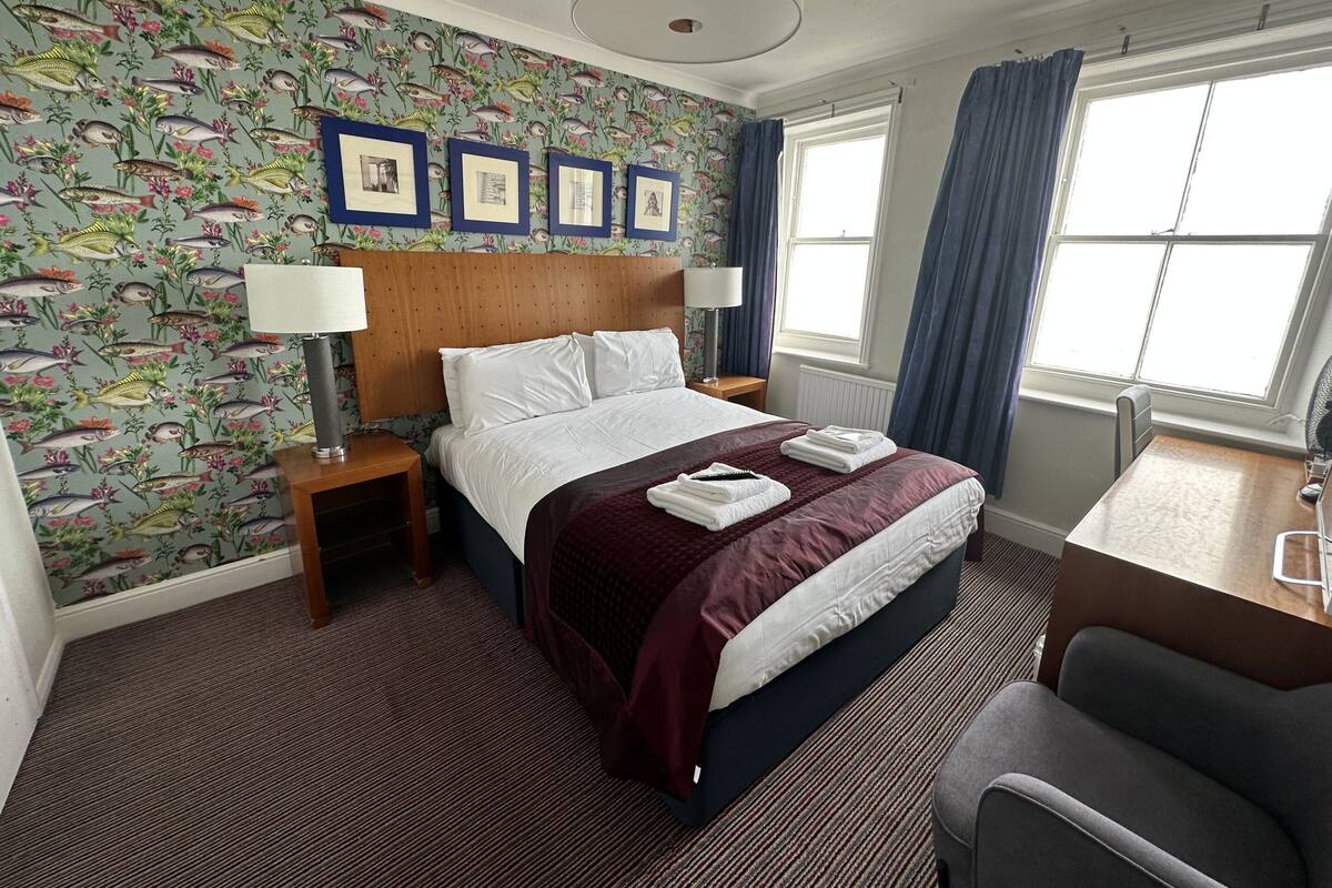 A hotel room with a bed, a desk, a chair, a window with curtains, and a wall with a fish pattern.