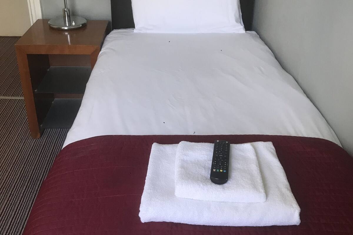 A hotel room with a neatly made bed, a remote control on a towel, a nightstand with a lamp, and a carpeted floor.