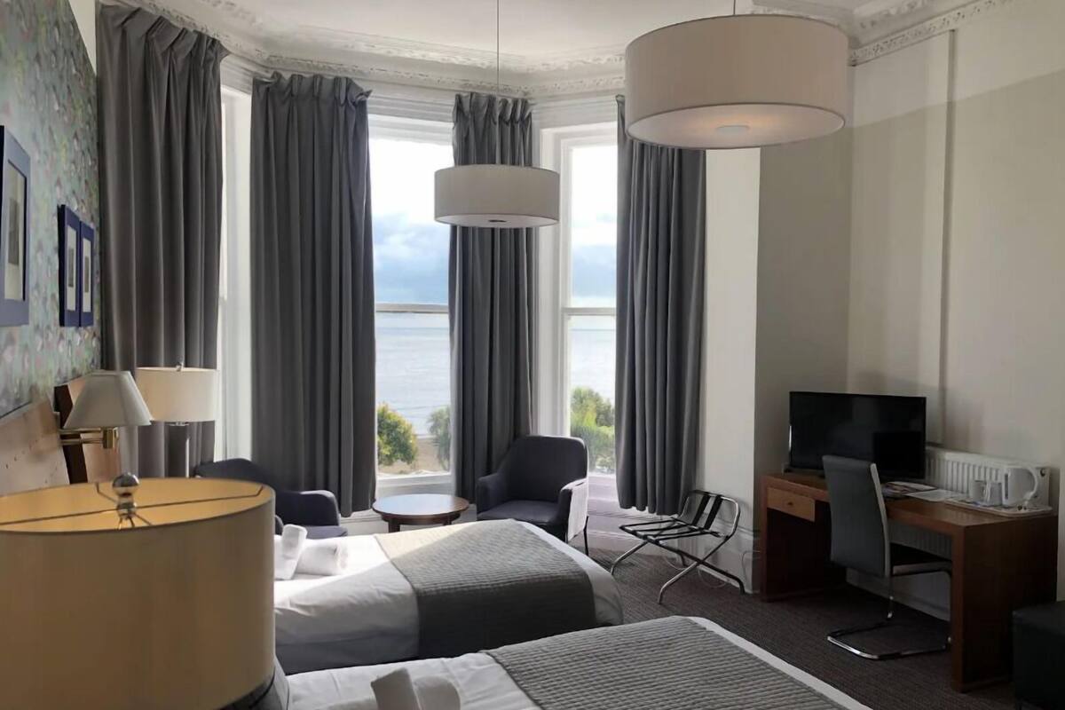 A hotel room with two beds, a desk, a chair, a TV, and a view of the sea.