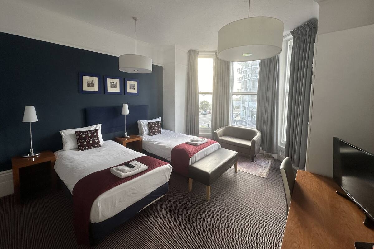 A hotel room with two beds, a bench, a TV, and a large window with curtains.