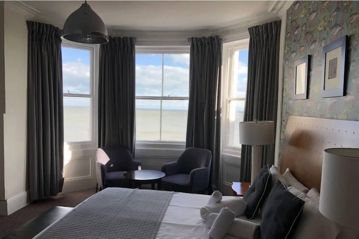A hotel room with a bed, two armchairs, a small table, a lamp, and a view of the beach.