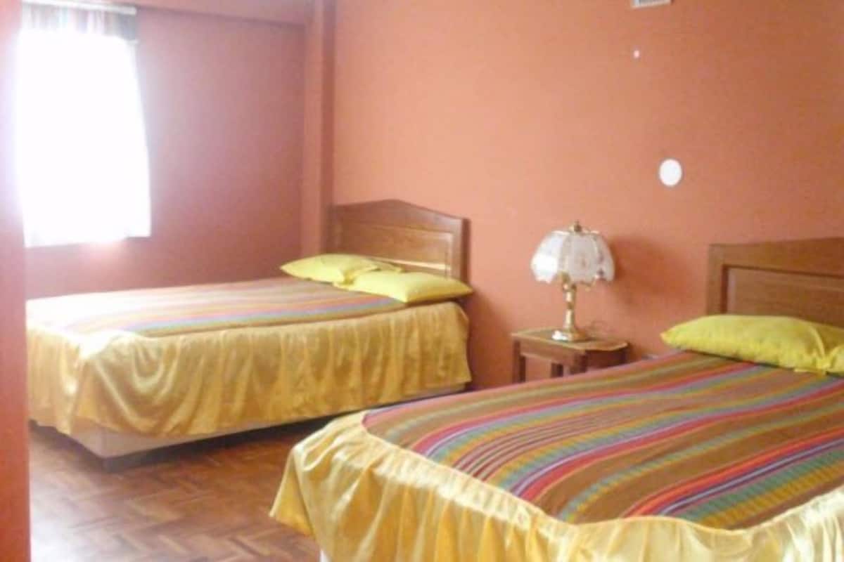 A room with two beds, each with a yellow bedspread and pillows, a bedside table with a lamp, and a window with curtains.
