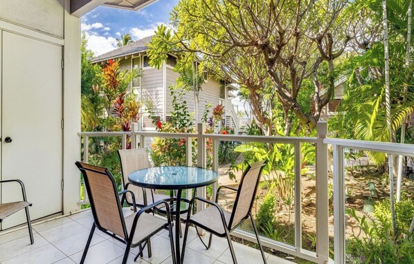 A balcony with a round table and four chairs, surrounded by a white railing, overlooking a garden with various plants and a building in the background. - Wailea Grand Champions (Wailea)