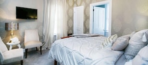 Ein Schlafzimmer mit Bett, Sessel, Nachttisch, Lampe und Fernseher an der Wand.