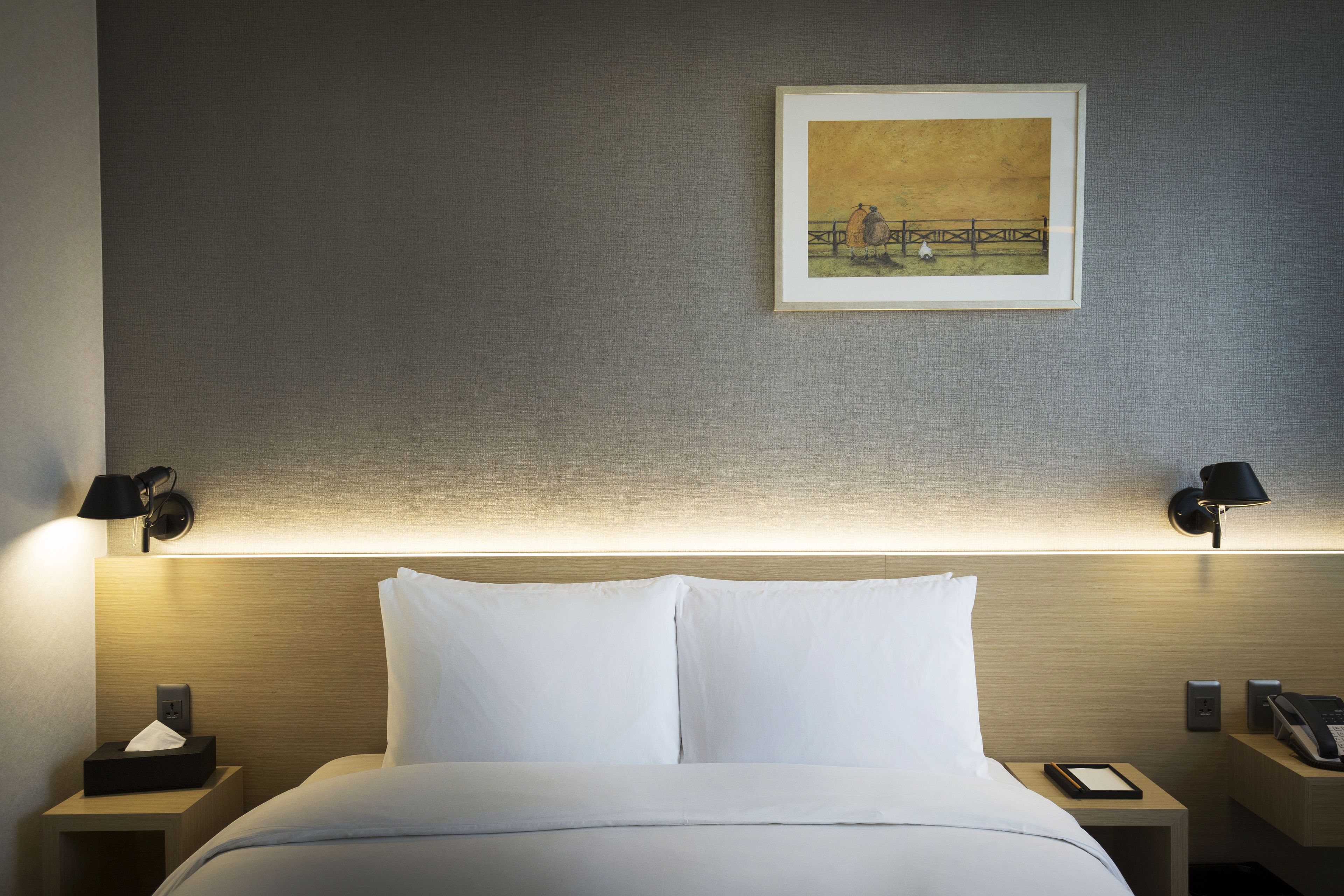 A neatly made bed with white linens, two bedside tables with lamps, a framed painting on the wall, and a bedside phone.