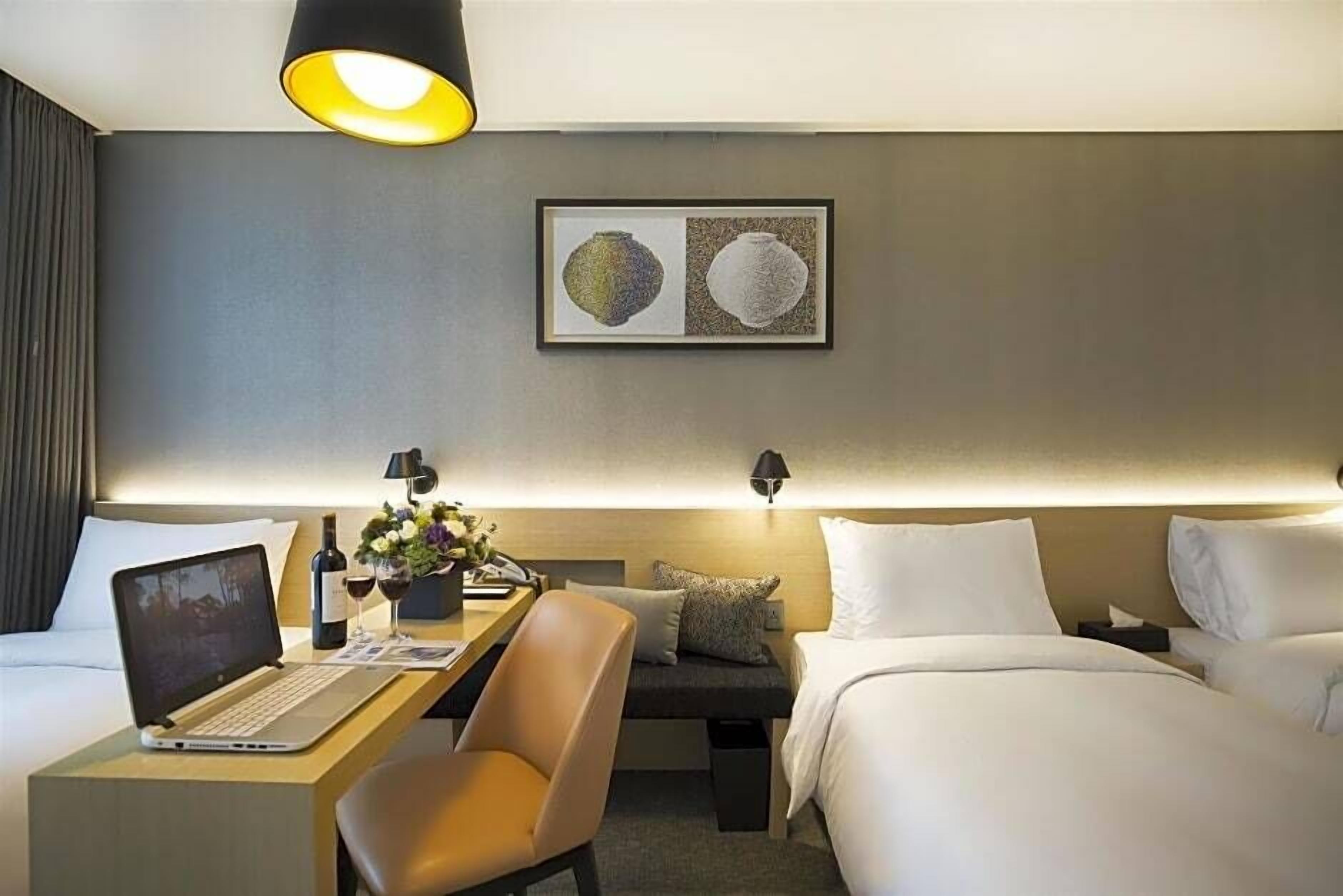 A hotel room with a bed, a desk with a laptop, a chair, a bedside table with a lamp, and a framed picture on the wall.