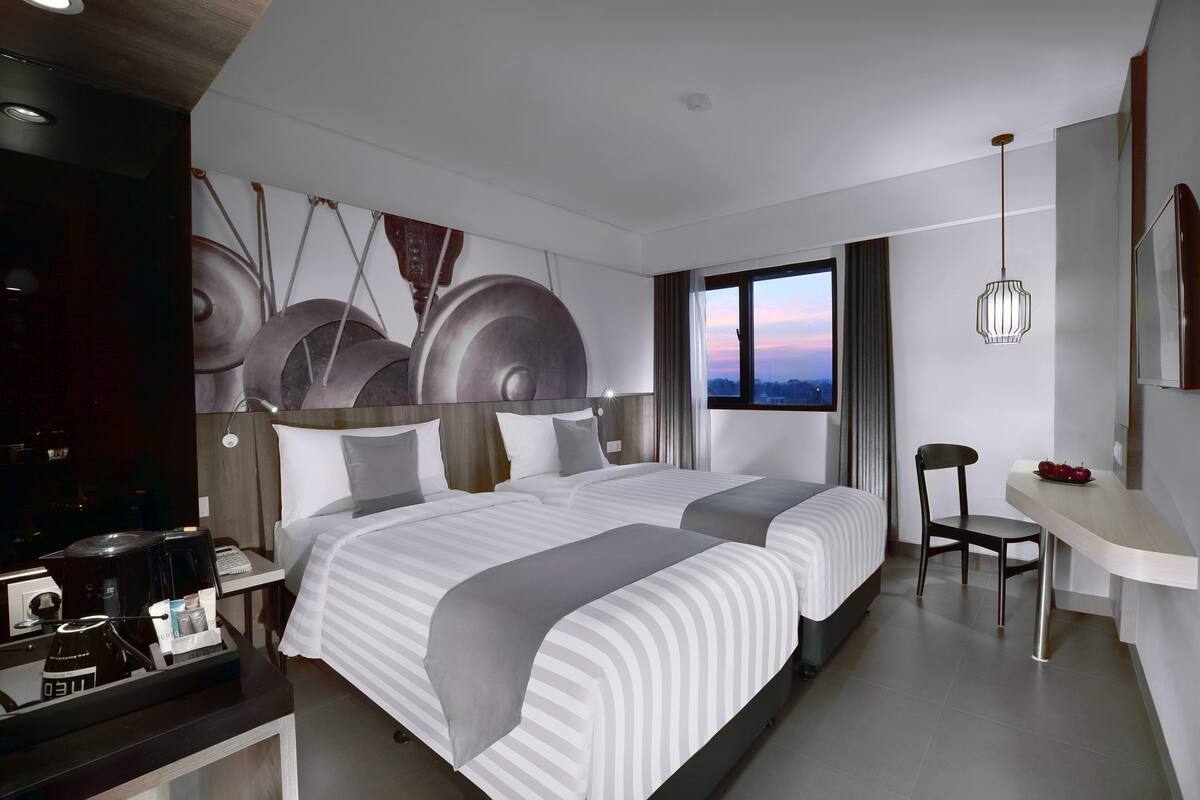 A modern hotel room with two beds, a desk, a chair, and a view of the sunset.