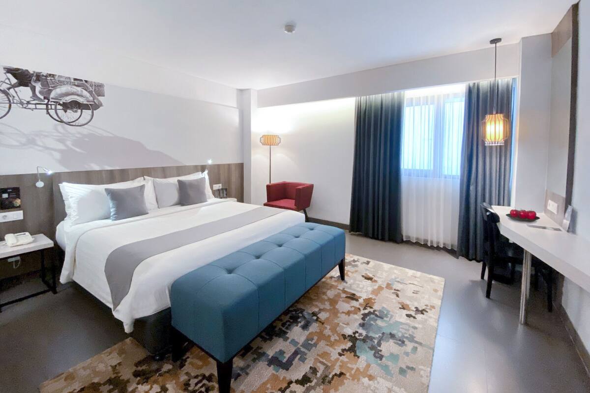 A modern hotel room with a large bed, a desk, a chair, a lamp, and a view of the outdoors.
