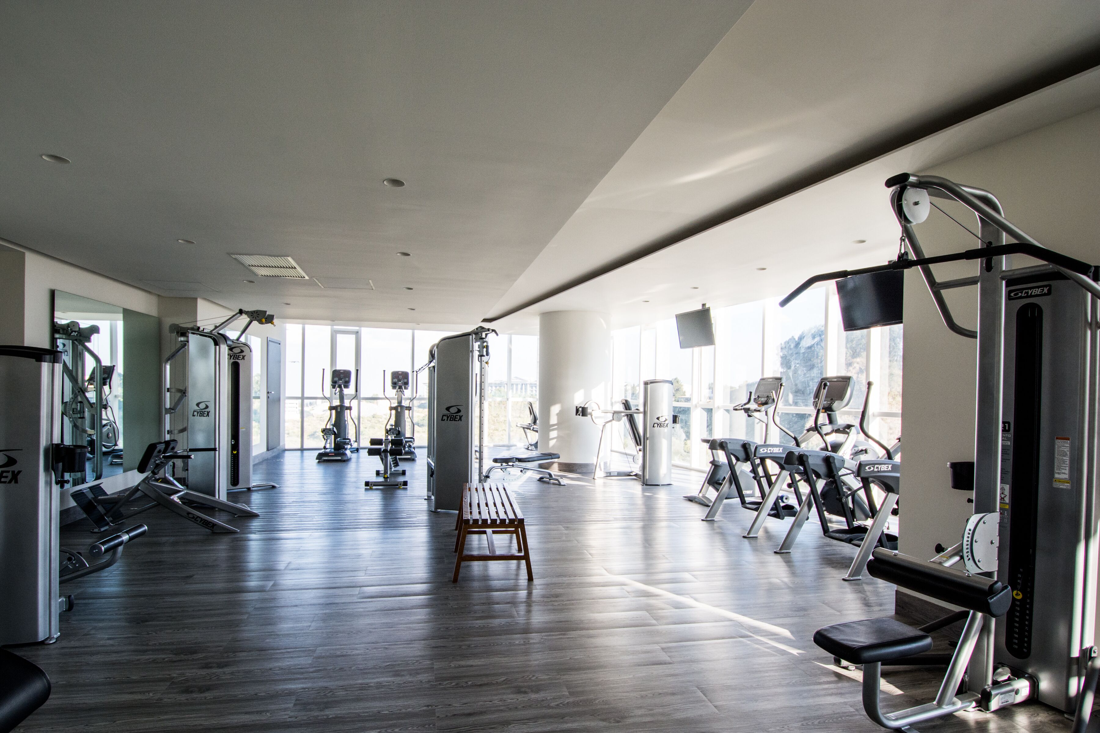A gym with various exercise equipment, a wooden bench, and large windows allowing natural light.