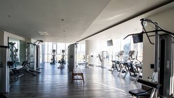 A gym with various exercise equipment, a wooden bench, and large windows allowing natural light.