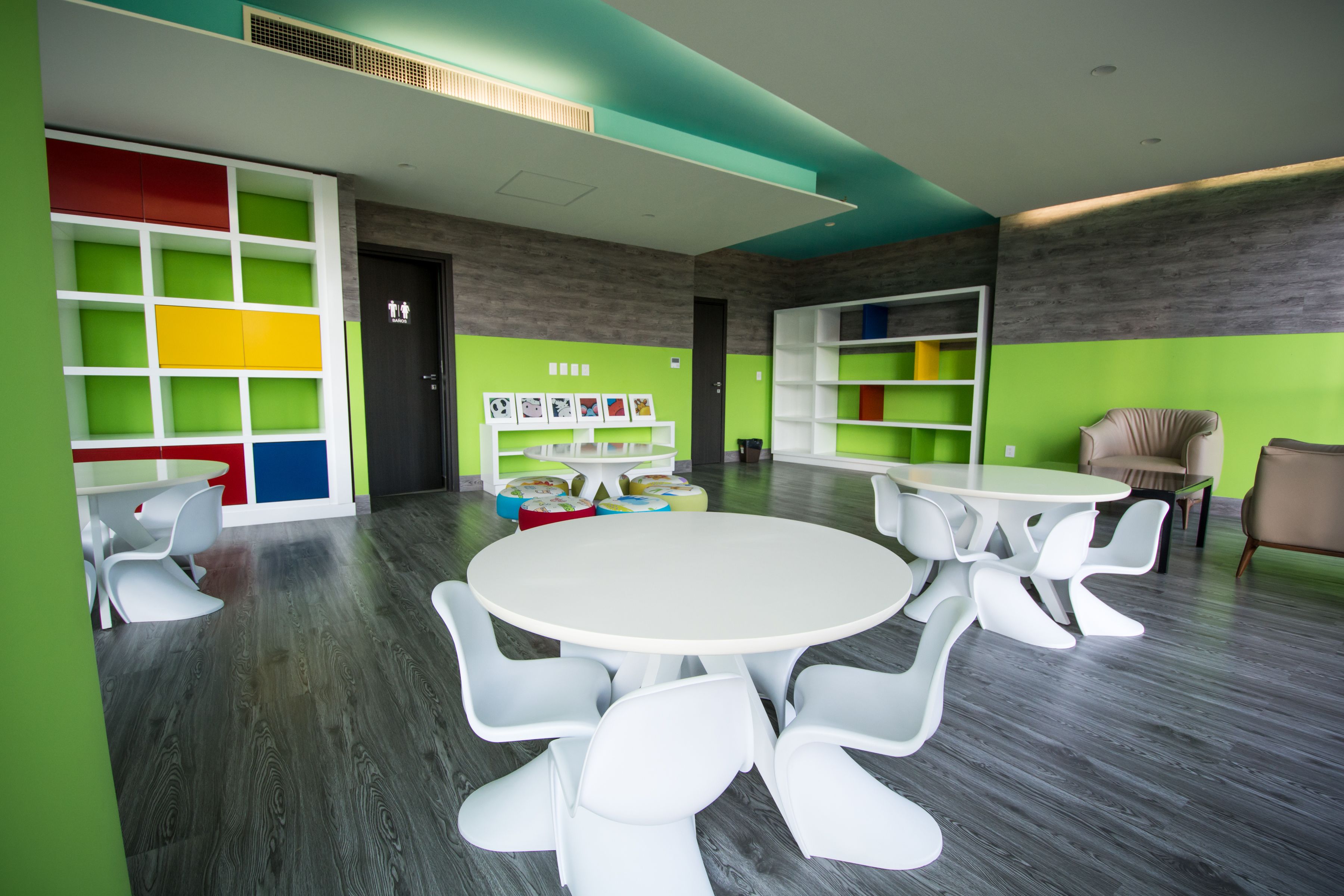 A modern children's playroom with white tables and chairs, colorful storage units, and a green and gray color scheme.