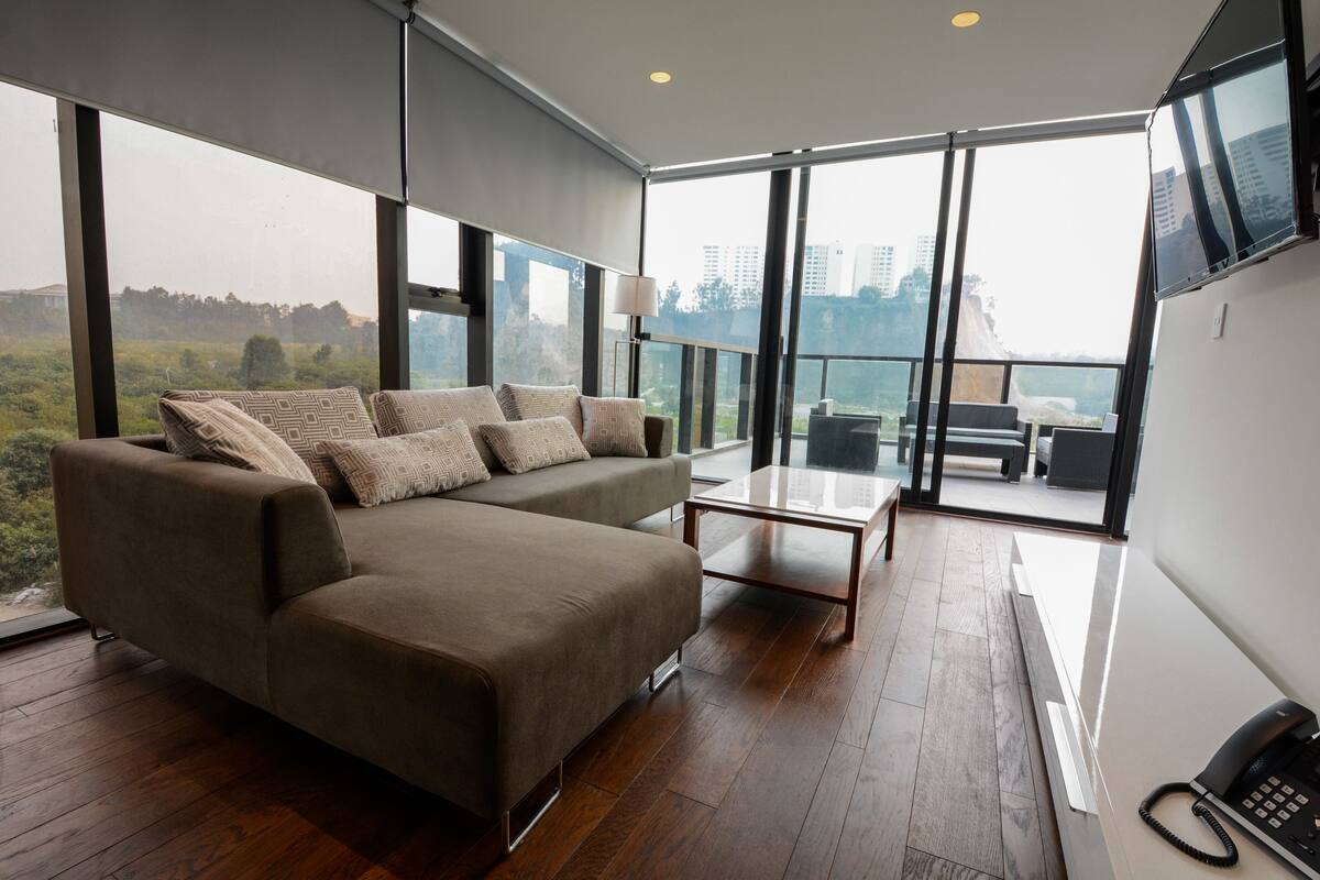 Ein modernes Wohnzimmer mit einer großen Couch, einem Couchtisch und Blick auf eine Stadtlandschaft durch große Fenster.