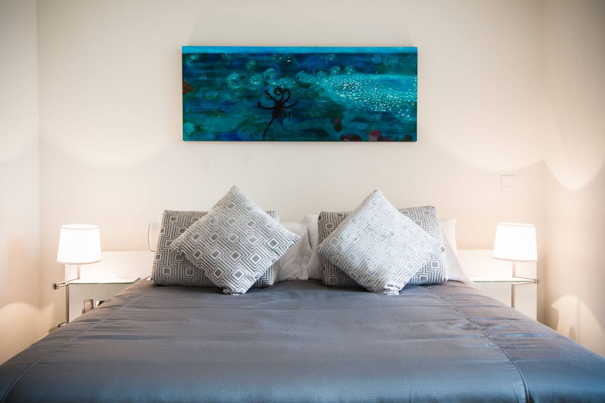 Ein ordentlich bezogenes Bett mit gemusterten Kissen, ein an der Wand hängendes Bild und Nachttischlampen.
