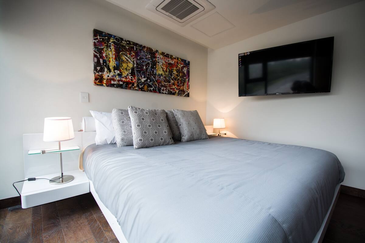 Ein modernes Schlafzimmer mit einem großen Bett, zwei Nachttischlampen, einem Flachbildfernseher und einem farbenfrohen, abstrakten Gemälde an der Wand.
