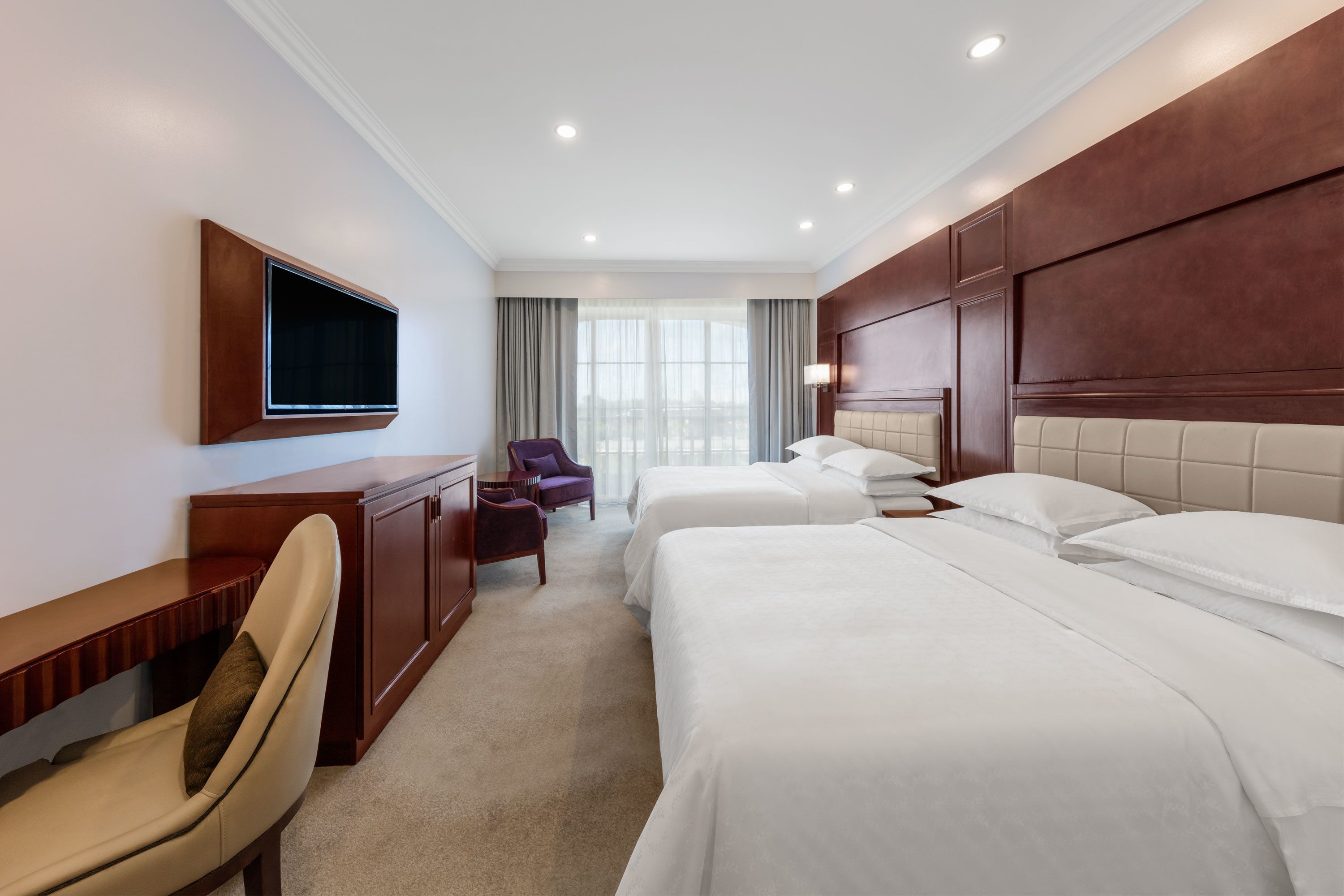 A hotel room with a large bed, a desk, a chair, a TV mounted on the wall, and a window with curtains.