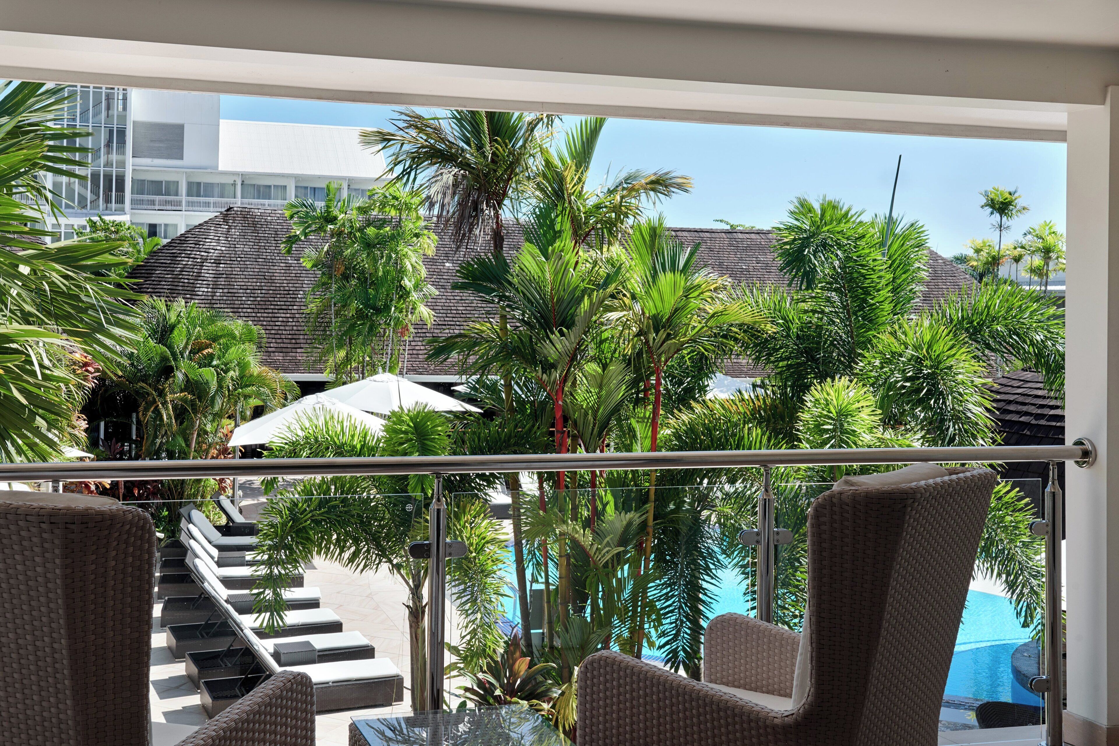 Un balcón con sillas de mimbre, una mesa de vidrio y vistas a una piscina y palmeras.