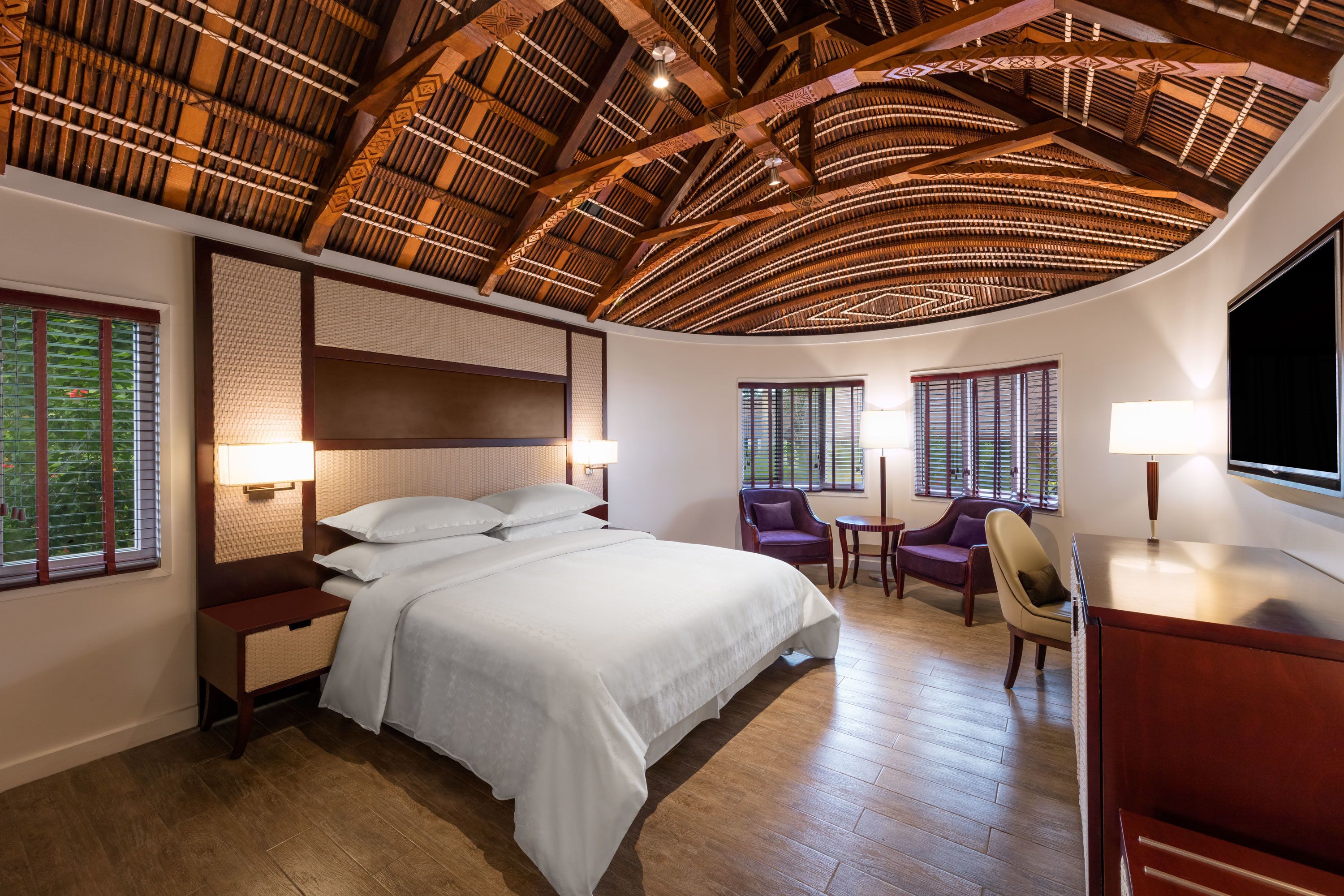 A hotel room with a large bed, wooden flooring, a high ceiling with a thatched design, two windows with blinds, and a television on the wall.
