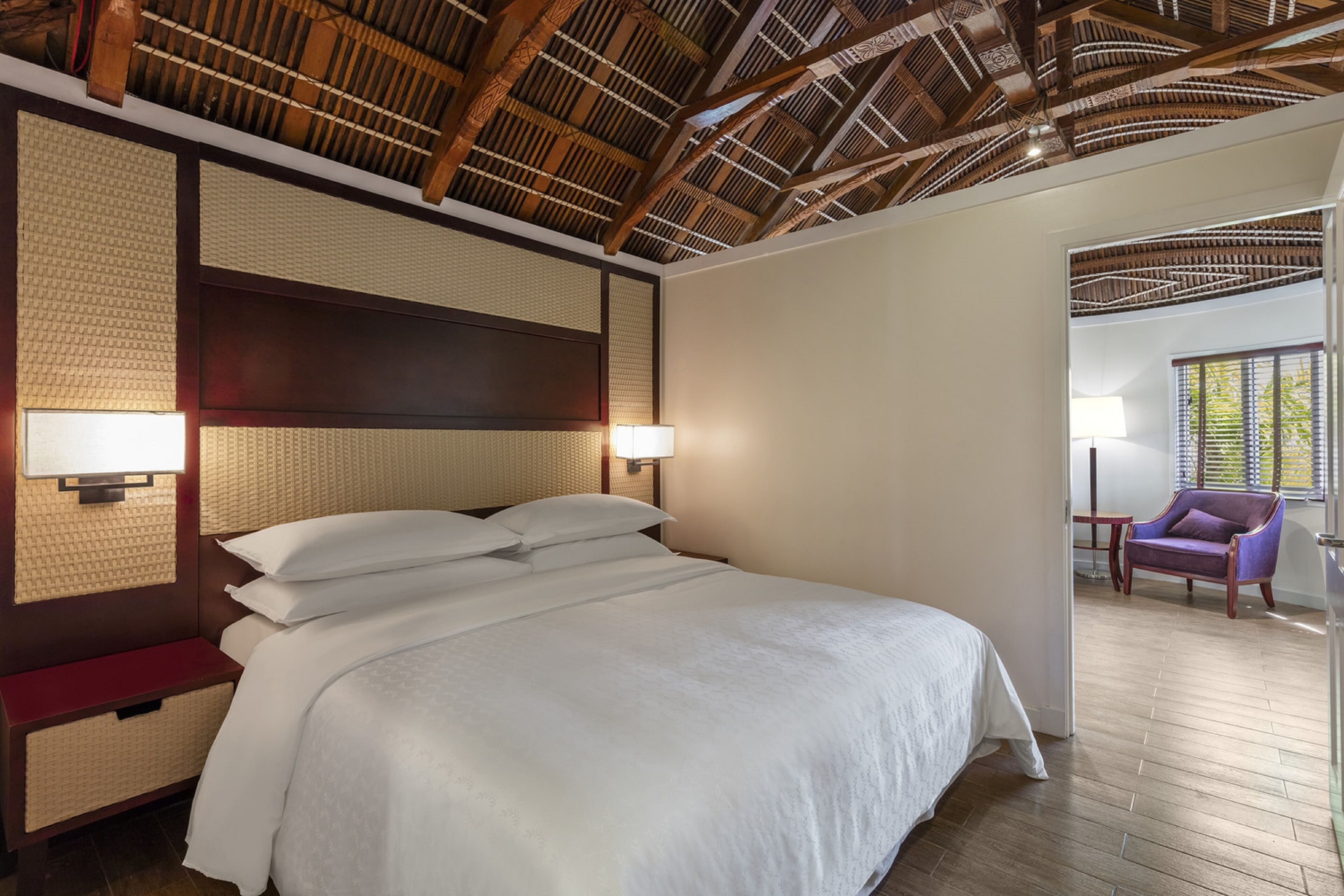 A hotel room with a large bed, wooden ceiling, and a chair by the window.
