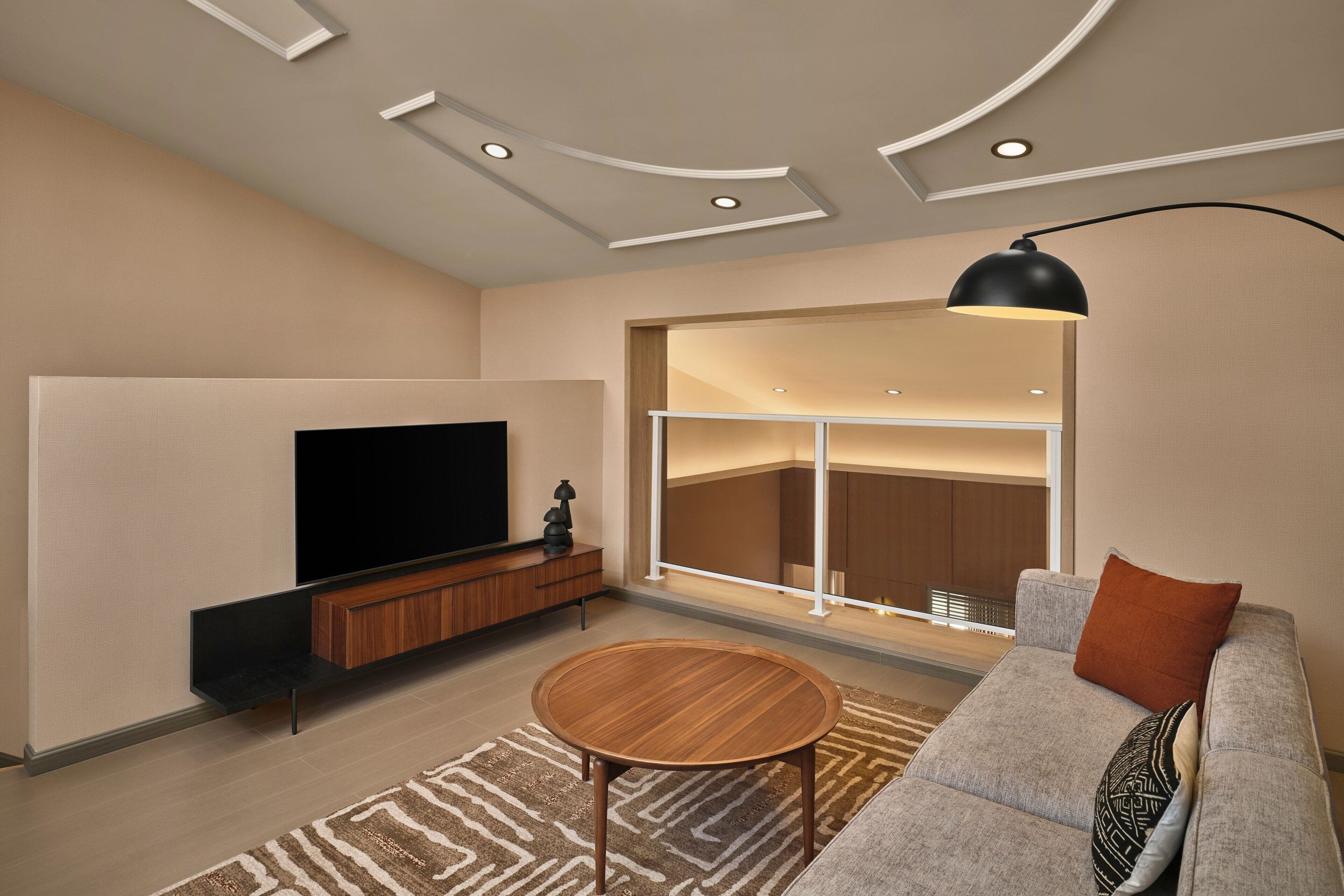 A modern living room with a flat-screen TV, a wooden coffee table, a grey sofa, and a large window with a view of the interior.