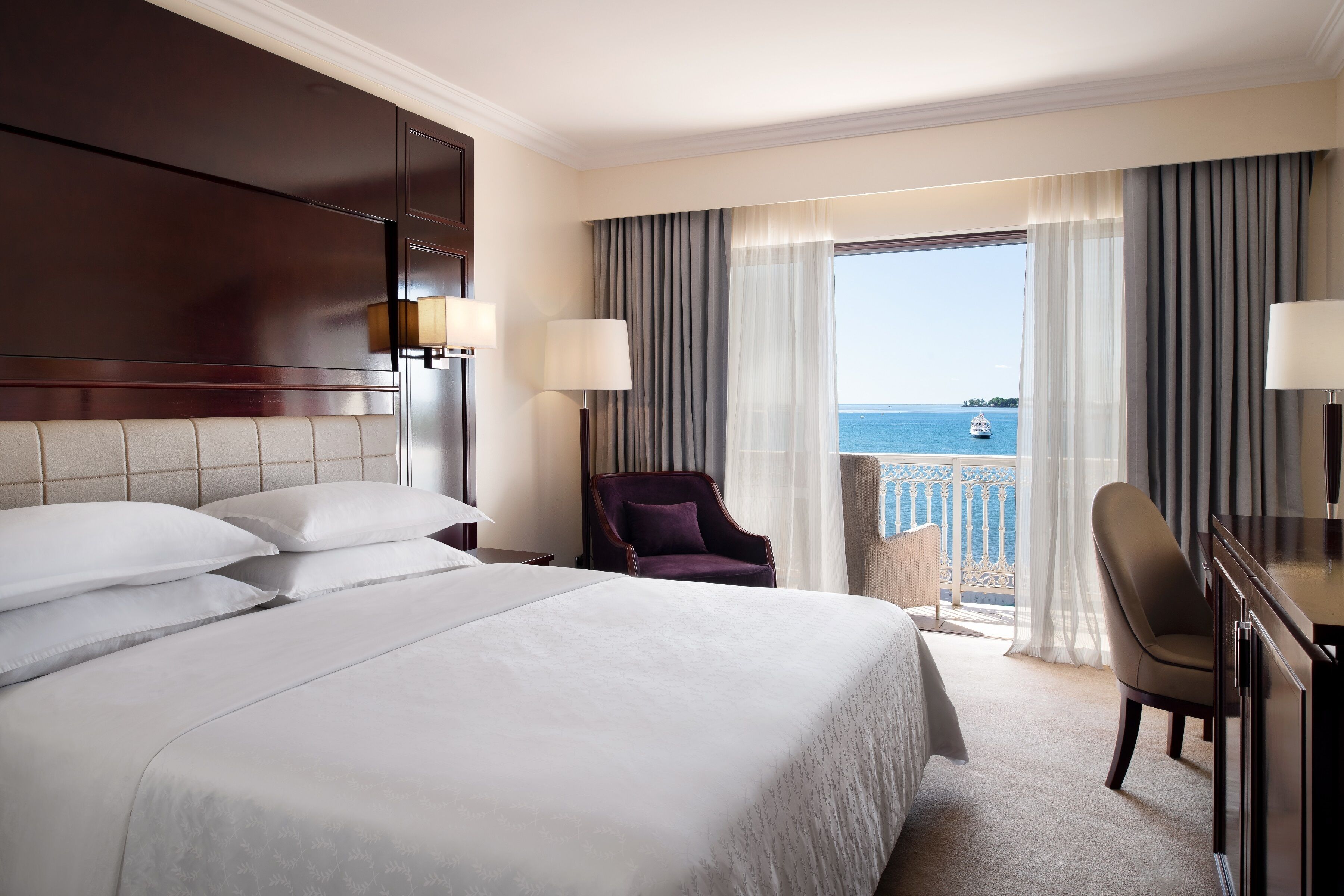 A hotel room with a large bed, two chairs, a desk, and a view of the sea.