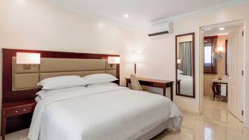 A hotel room with a large bed, bedside tables, a desk, a chair, a mirror, and a bathroom visible through an open door.