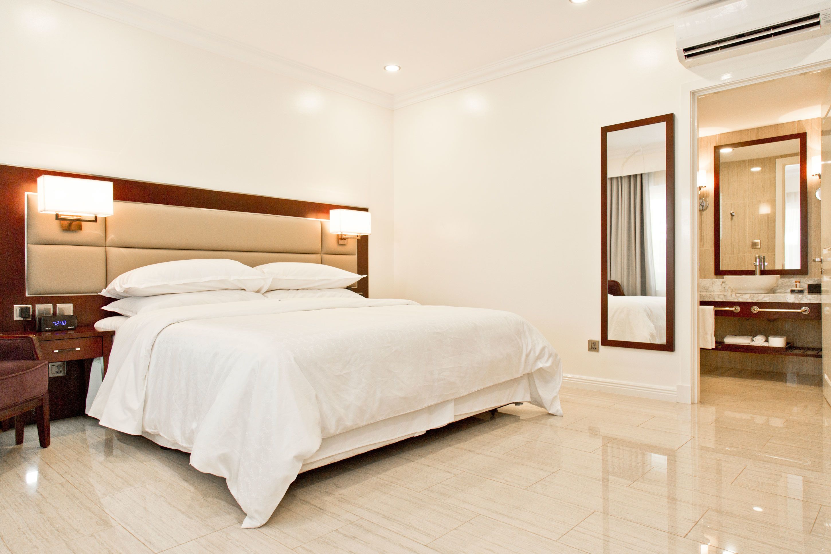 Habitación de hotel con una cama grande, lámparas de noche, mesita de noche, una silla, y un baño con espejo, lavamanos y toallero.
