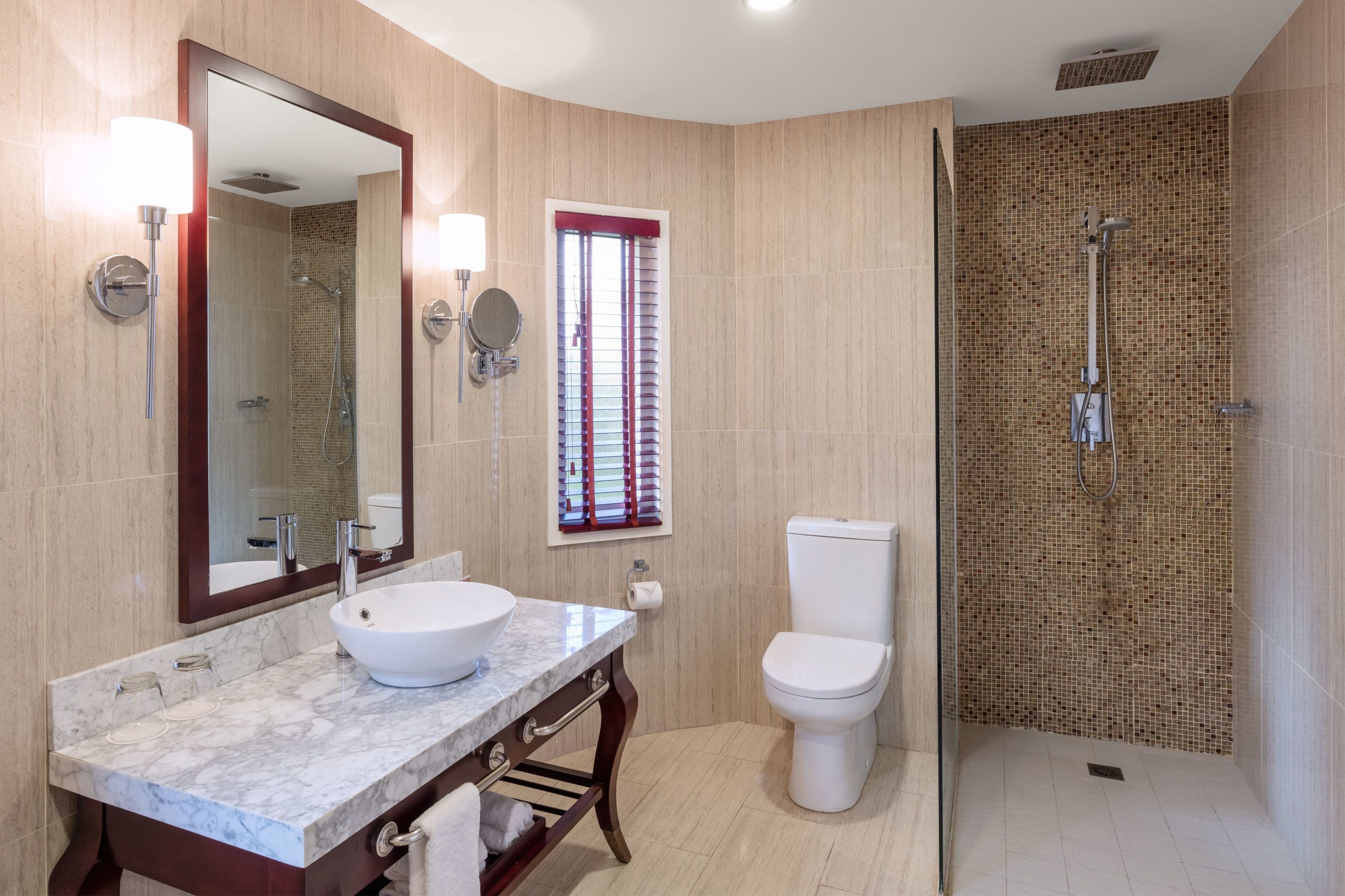 A bathroom with a marble sink, a toilet, a shower with a tiled wall, and a mirror with wall sconces.