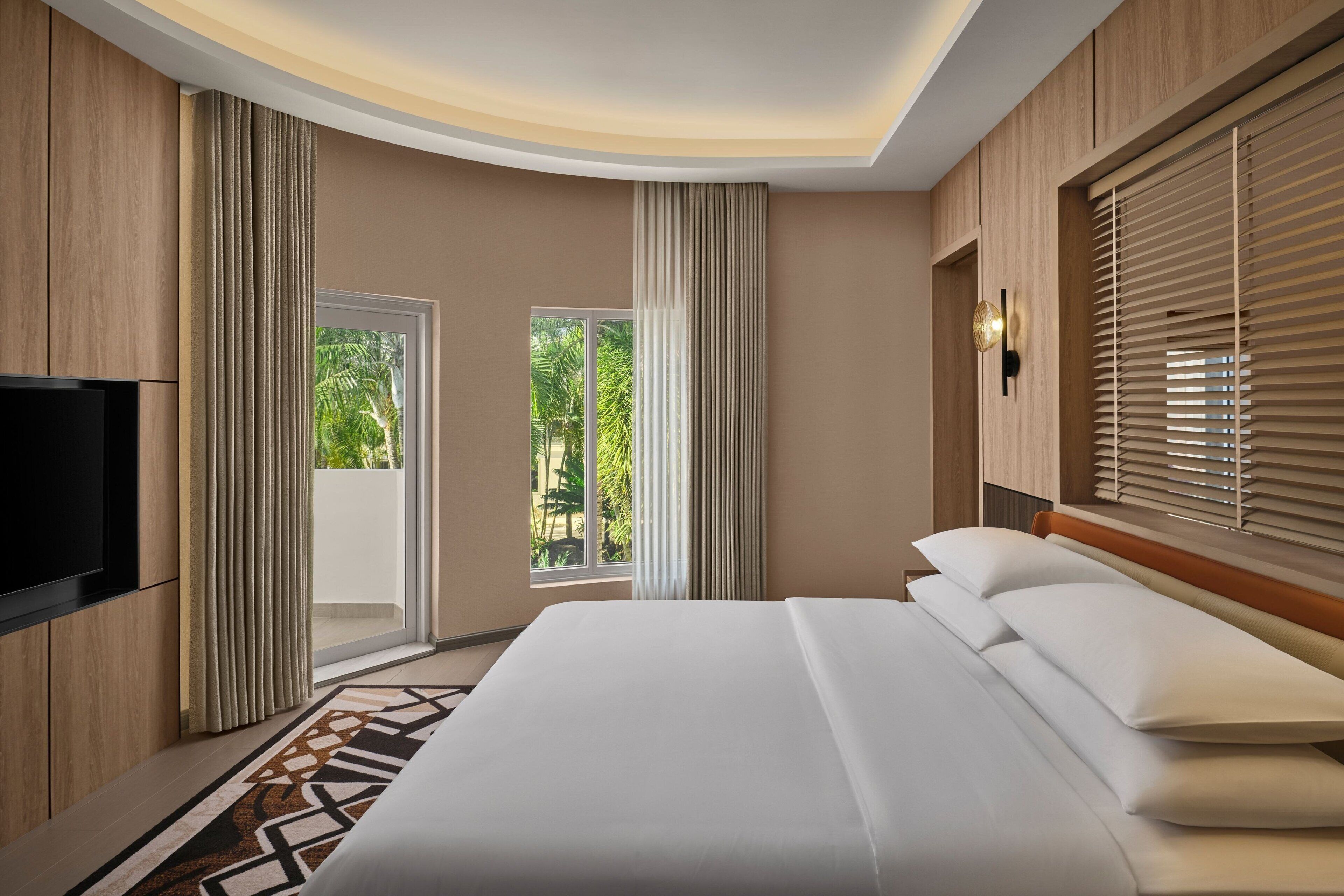 A hotel room with a large bed, a flat-screen TV, and a sliding glass door leading to a balcony with a view of greenery.