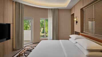 A hotel room with a large bed, a flat-screen TV, and a sliding glass door leading to a balcony with a view of greenery.