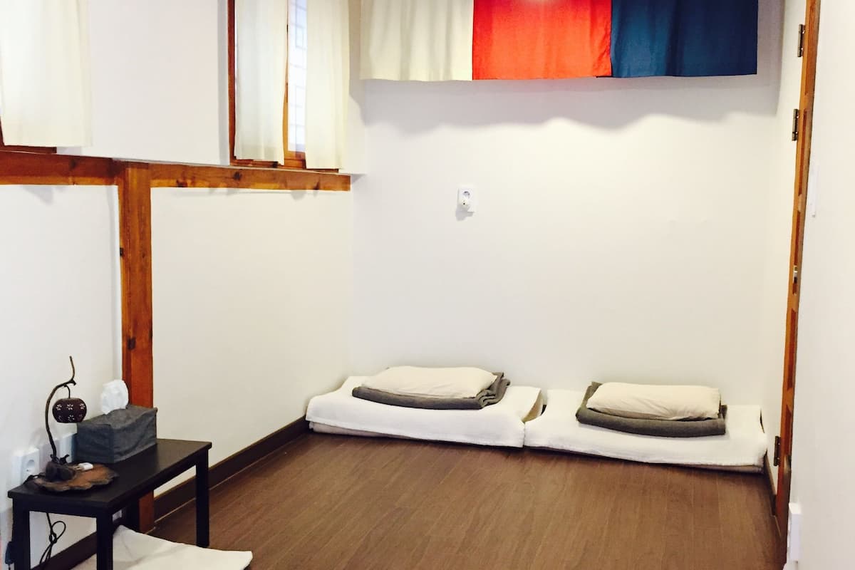 A room with wooden beams, a bed with pillows, a small table with a lamp, and a window with curtains.