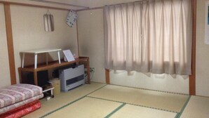 Una habitación pequeña de estilo japonés tradicional con suelo de tatami, un futón, un escritorio de madera bajo y un gran ventanal con cortinas.