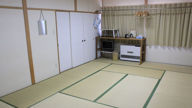 Una habitación con suelo de tatami, una futón, un escritorio pequeño con un ordenador y una ventana con cortinas.