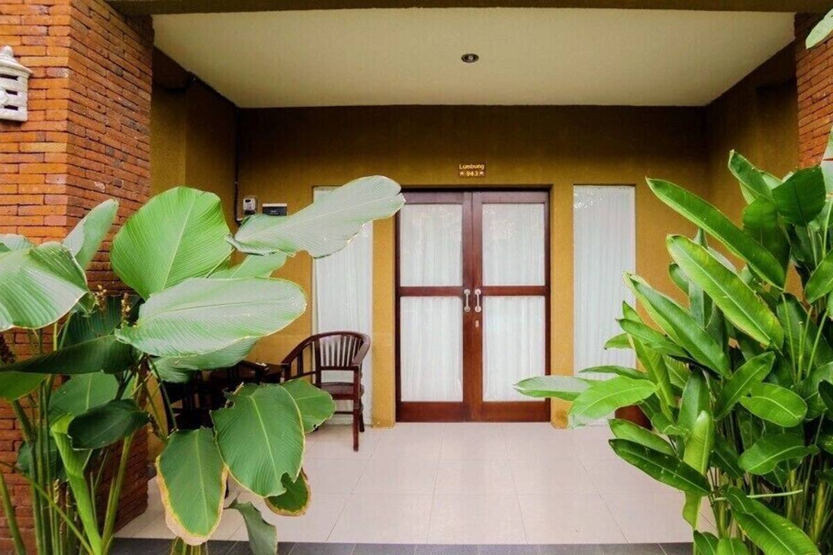 A covered entrance with a wooden door, a chair, and large green plants.