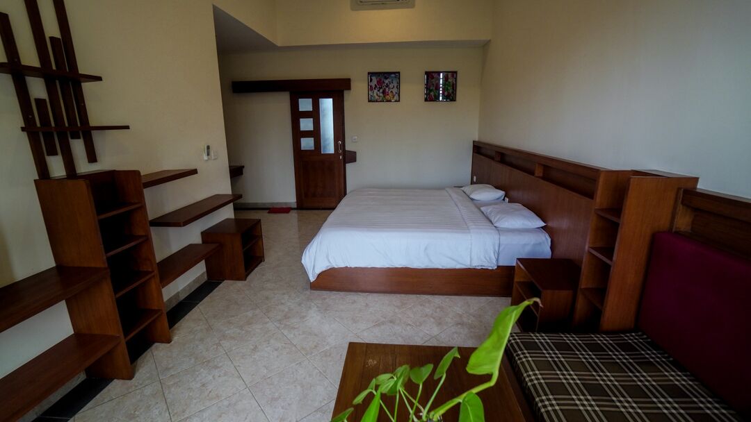A bedroom with a large bed, wooden headboard, a wooden bench, a small table with a plant, and a door leading to another room.